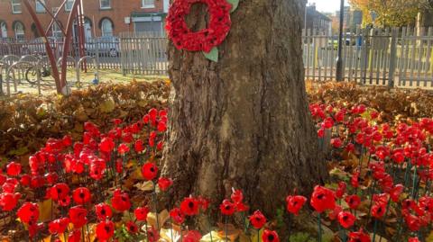 Red plastic poppies on sticks around the base of a tree, which is surrounded by brown autumn leaves. There is a metal grey fence and houses behind and bike racks to the left