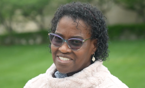 A woman with short, dark curly hair talks to the camera. She wears sunglasses and a cream jumper.