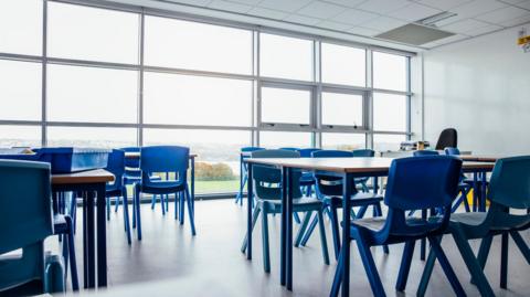 A generic image of a classroom - there are several desks with classroom chairs and a window in the background with a view of the countryside in the background.