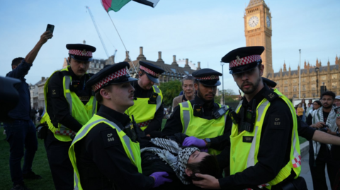 Police officers remove a protester as demonstrators take part in a Lift the Ban on Palestine Action protest organised by Defend our Juries in Parliament Square in London. There are five officers in hi-vis vests and police hats and one is cradling the protester's head.