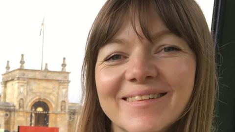 A woman with long straight brown hair smiles with an historic-looking building in the background. 