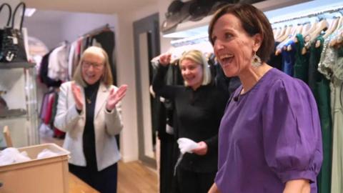 Anne is in the foreground wearing a bright purple blouse and speaking with an animated smile to someone opposite. They are in a dress shop, and two women behind her are clapping and celebrating.