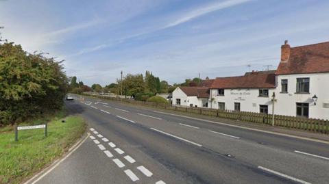 View of a road, with a white pub with a red roof on one side of the road, and grass and a road sign on the other