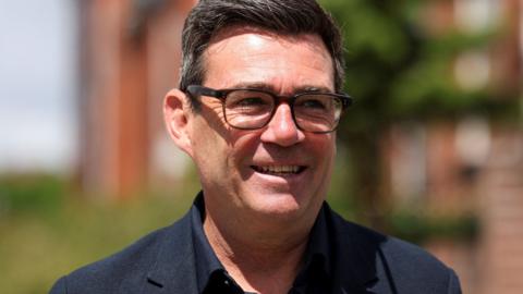 Greater Manchester mayor and Labour candidate for the Makerfield by-election Andy Burnham. He has dark brown hair with some grey wearing black glasses and a navy top and blazer. He is standing in a street which is blurred out. He is smiling.