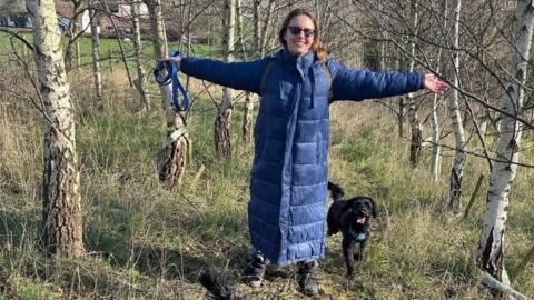 Adele Hawkins-Waterfall with her dog. She is wearing a long blue coat and is wearing glasses. She has her arms outstretched and she is surrounded by trees.