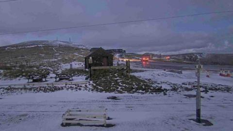 A view of the A18 Mountain Road at the Bungalow, which remains snowy, but the rear lights of vehicles can be seen travelling on it.