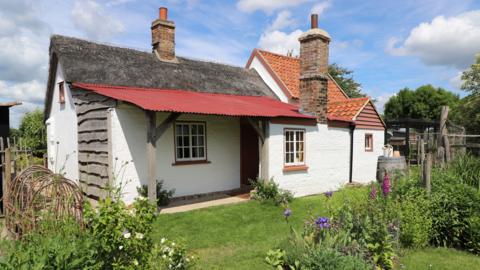 A small single storey cottage is located in a garden which has several flowers and trees. One side of the building is thatched and the other has red roofing tiles.