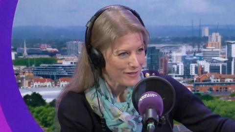 Donna Jones sits in the BBC Radio Solent studio with a backdrop of Southampton behind her. She has long blonde hair with headphones on and a navy blue knit jumper and silk floral scarf round her neck.