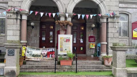 The outside of a church building decorated with bunting and a sign for the immersive arts installation. Grass and steps in front of the two panelled door building.