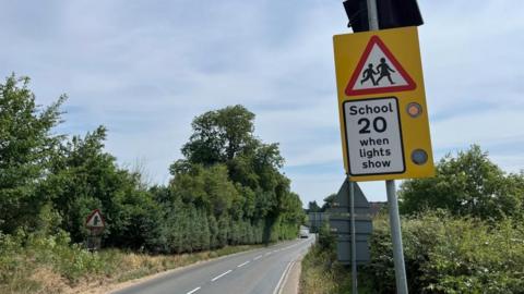 A warning sign, in yellow, featuring a warning triangle depicting two children crossing the road, and a segment stating: "school 20 when lights show". One of the two lights, which alternately flash, is illuminated.