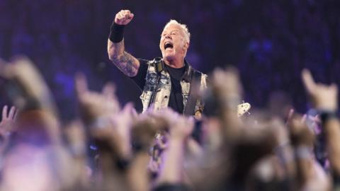 Lead singer and guitarist of Metallica, James Hetfield performs onstage at Lumen Field. He's surrounded by a crowd of people with their arms up. He's wearing a black t shirt, arm cuff and a denim vest. He's holding a guitar. He's got his arm up in the air and appears to be shouting.