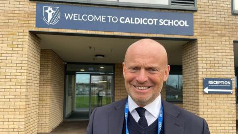 Alun Ebenezer wears a suit and a lanyard, smiling at the camera in front of a sign saying Welcome to Caldicot school