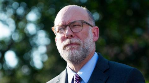 Russell Vought speaks to media outside wearing his glasses, a striped tie, button-down shirt and dark suit jacket
