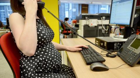 A  woman in a black and white polka dot dress sitting at a desk in an office. She has one hand on the computer keyboard and the other is holding the desk phone receiver.