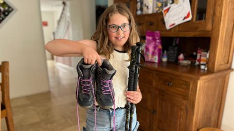 A young girl stands in a rural kitchen holding a pair of hiking boots and some walking sticks.