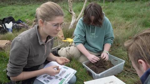 Head and shoulders shot of two student looking at a soil sample in a Sussex meadow. One student poring through the mud and the other has a worm and a worm identification sheet.