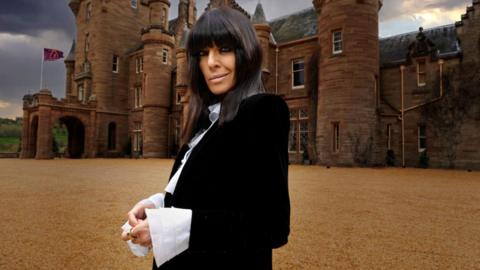 Claudia Winkleman stands side-on looking down at the camera with a conspiratorial smirk. She is wearing a black jacket and white shirt with big cuffs and her trademark long fringe. Ardross Castle stands moodily behind her with a Traitors flag flying on the flagpole.