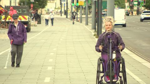 Terri Ballon walks along the Strand using a white cane to guide her. On the right Dr Kay Hinkle rides her arm-pedal bike 