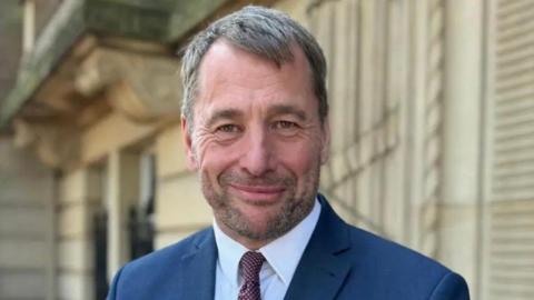 Councillor Stephen Atkinson standing outside County Hall in Preston smiling. He has short fair hair and stubble and is wearing a red rose badge on his navy blue suit jacket.