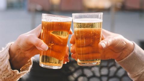 A close-up of the hands of two people clinking pin glasses of amber-coloured beer together in front of an out-of-focus background.