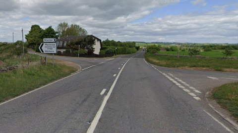 Junction of the A92. There is countryside around the road and a house and sign. 