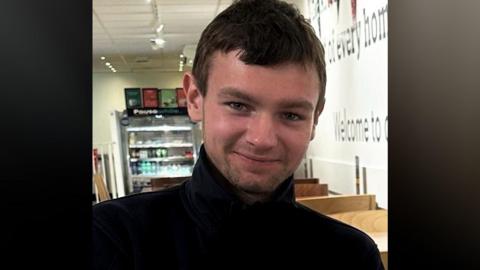A man wearing a black round-necked top. He has short, brown hair and stubble and he is smiling.