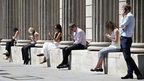 Five people sit along a low wall in the sunshine looking at their mobile phones, while a man stands at the right-hand side speaking to someone on the phone