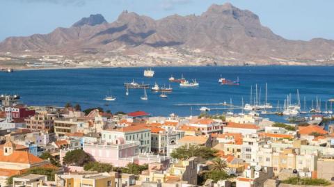 View over Mindelo, Sao Vicente, Cape Verde showing buildings with terracotta-coloured roofs in front of the blue ocean, which has several boats in it. Behind the ocean is a grey mountain and the sky is light and blue.