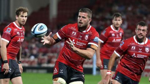 Ed Slater wearing a red football shirt with black shorts, catching a rugby ball while running on the pitch alongside three other players from his team. It is captured before his retirement following his MND diagnosis.