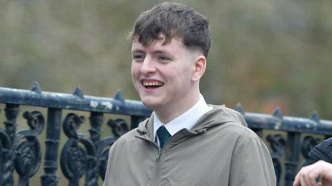 Ciaran Wason wearing a shirt, tie and grey/green jacket. He has short, brown hair and is smiling.