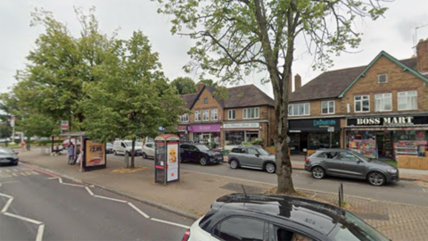 General view of a row of shops on Stratford Road, Shirley. The line of terrace shops is on the other side of the road with cars parked outside.