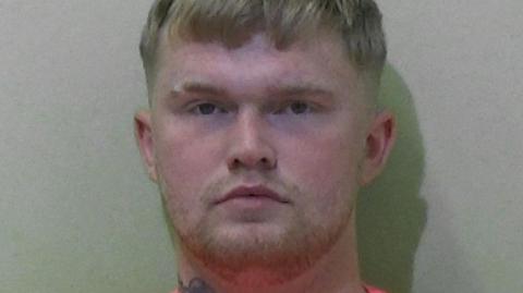 Mugshot of Caris. He has faint blond stubble and short blond hair.