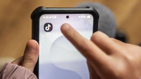 In the foreground is a child's finger reaching towards a TikTok logo on a phone screen. The phone is being held by their other hand. The background is out of focus but wooden flooring and dark coloured trousers can be made out.