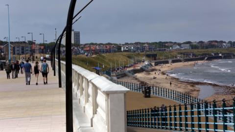 The promenade and coastline in Whitley Bay. People are strolling along the left-hand side of the picture, while the beach appears to the right.