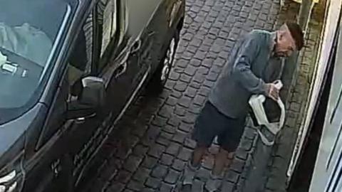 CCTV footage showing Maughan affixing a black box to a bollard using white tape. The bollard is outside a shop. Maughan is wearing shorts and has short hair which has been shaved completely around the back and sides of his head. He is standing behind a black van.