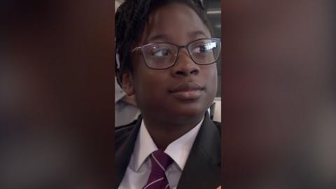 A young girl wearing school uniform, a white shirt and a black blazer. She has dark glasses.