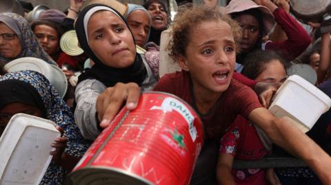 A large crowd of people—adults and children in Gaza—gathered under a makeshift shelter made of tarps. Many are reaching out with empty containers like pots, pans, and buckets, their faces marked by urgency and desperation.