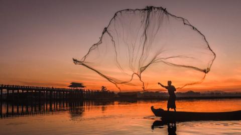 Silhouette of a man casting a large fishing net in the air from a boat during sunrise