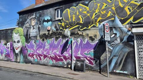 A large street art mural painting on the side of an electronics store in Swindon. It shows a dark city landscape with the bat signal cast in the sky above, a blue and black batman with his mask and cape, the Joker and The Penguin. It is a bright sunny day and the sky is blue.