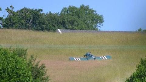 Field surrounded by trees with a light aircraft in the grass.