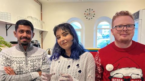 Three young people look at the camera. Naveed (left) is Asian heritage and had a beard. He wears a grey and white jumper with a snowflake pattern. Anya wears a holey white jumper with a black shirt underneath. She has blue/black hair, dark eyeliner, a nose piercing and is holding two glasses. Kamil has ginger hair and glasses and is wearing a Christmas jumper. 