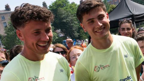 Two men in yellow T-shirts with logos on. They are talking to a reporter who is out of shot. 