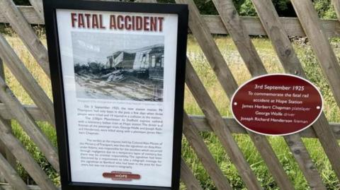 Commemorative plague with a black frame and a grass shiny cover contains the story about a rail disaster at Hope Station in Derbyshire. There is a black and white image at the top which shows a train carriage destroyed by the impact of the head-on crash at Hope Station 100 years ago. The heading is in red and reads "Fatal Accident". The type face is in black and tells the history of the disaster, which claimed three lives. The plaque is attached to a wooden fence which has diagonal slays. Next to it is an overal-shaped plague, which reads "3 September 2025" to commorate the fatal rail accident at Hope Station and names the three that died. The writing is in which and set on a red background. 