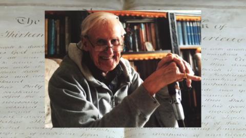 A smiling elderly man in glasses with a walking stick sits in front of shelves stacked with old books