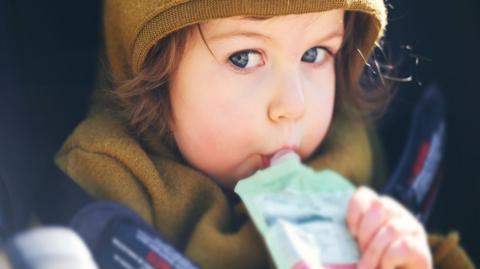 A stock image of a young child with brown hair sucking on a pouch of food
