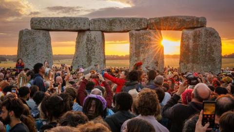 Thousands of people gathered in a crowd - many holding up cameras and phones - watching the sun rise up behind the stones at Stonehenge, with the silhouettes of trees and a yellow, cloudy sky in the background. 
