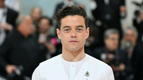 Rami Malek has short black hair and is wearing a white vest and shirt with a green broach. Behind him, there are photographers out of focus. 