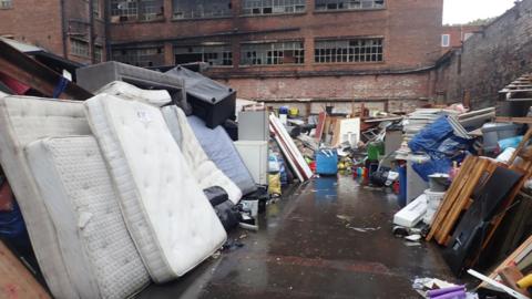 An outdoor yard filled with large quantities of discarded waste and bulky household items sits in front of a derelict multi-storey red-brick industrial building. In the foreground, several stained mattresses are stacked upright alongside upholstered furniture, cabinets, shelving, timber, plastic containers, and black rubbish bags.