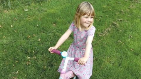 Alysia Salisbury is a blonde little girl wearing a mauve summer dress and sitting on a pink Barbie bicycle. She has a big smile on her face. 