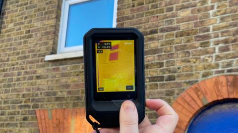 A thermal imaging camera being held up towards the top left window of a house. On the screen it shows yellow on the bricks, and the window is red. 